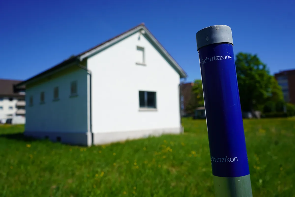 Heute weisen blaue Pfosten auf die Schutzzone hin. Künftig wird das Häuschen eingezäunt sein. Tinkwasserfassung Feld, weisses Häuschen steht auf einer grünen Wiese